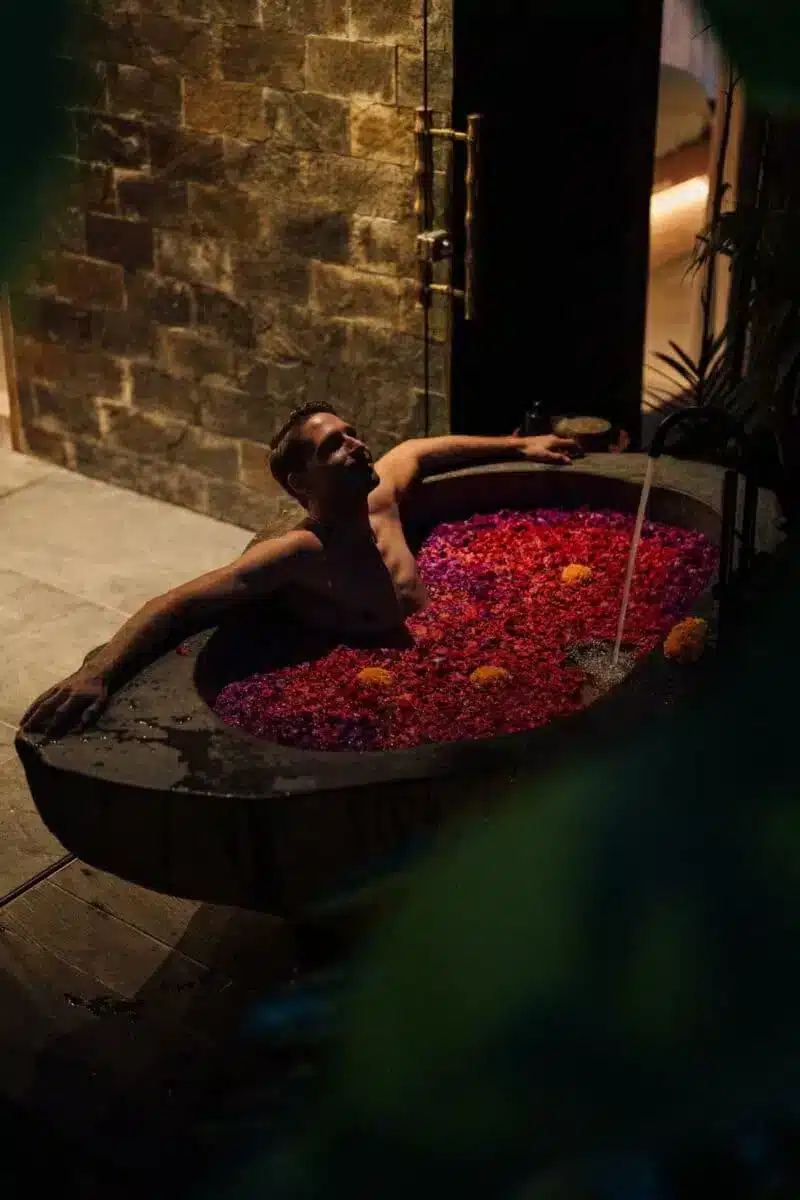 Guest enjoying a traditional Balinese flower bath filled with red and pink petals in the outdoor stone bathtub at Desa Hay Bali