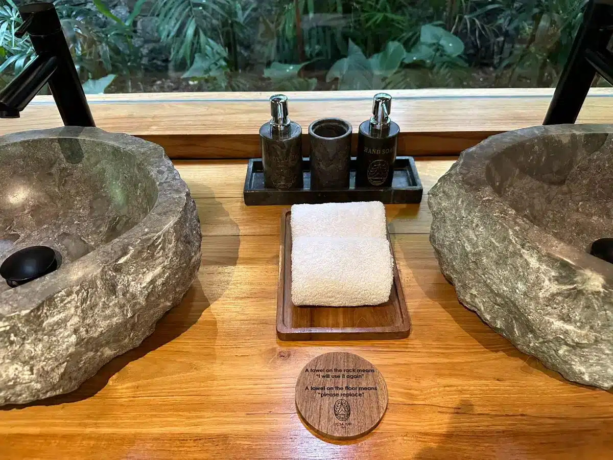 Bathroom amenities including natural stone basin toiletries tray and folded towels on wooden counter at Desa Hay Bali