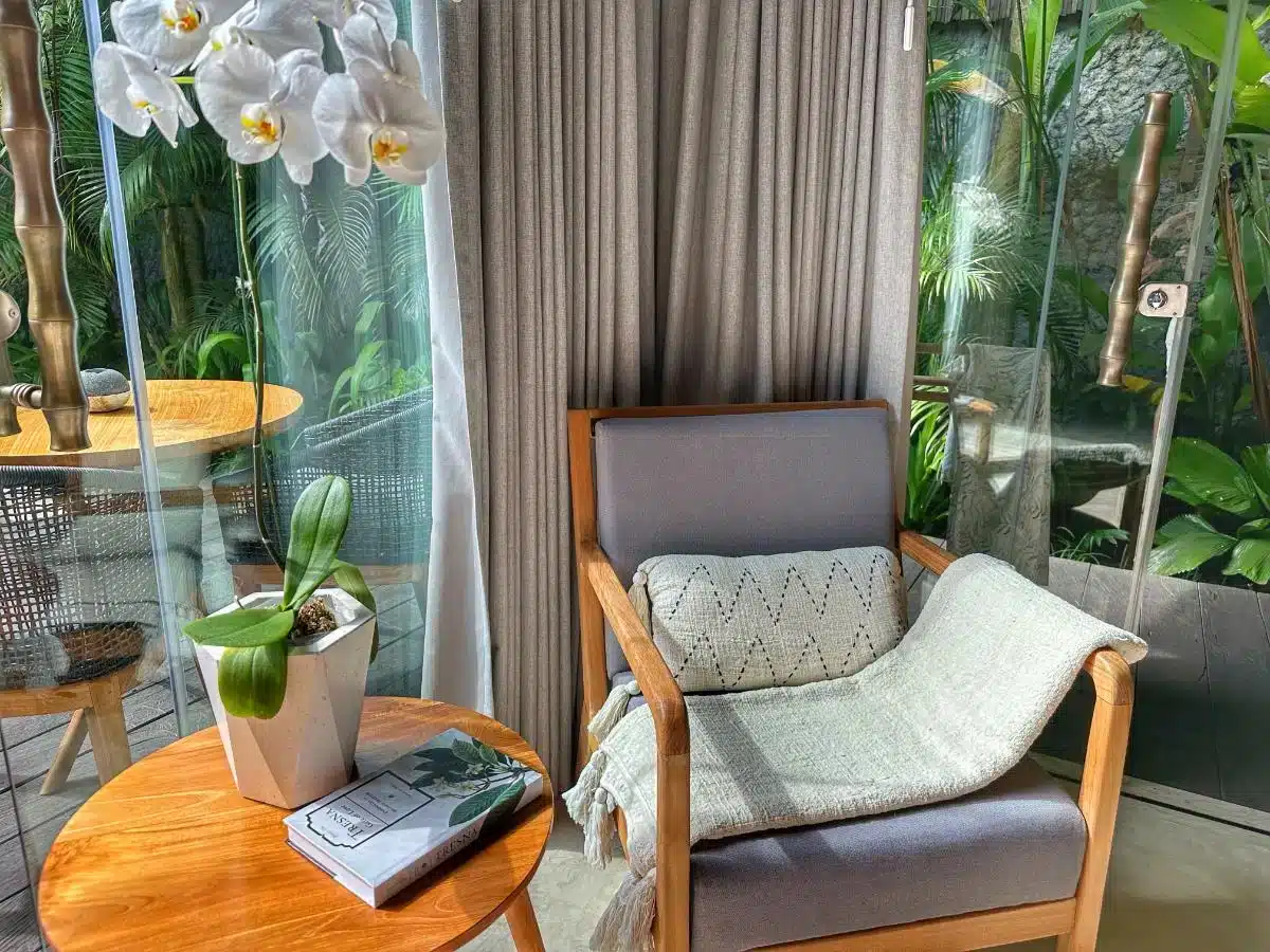 Villa seating area with armchair orchid arrangement and side table next to floor-to-ceiling glass windows overlooking tropical garden