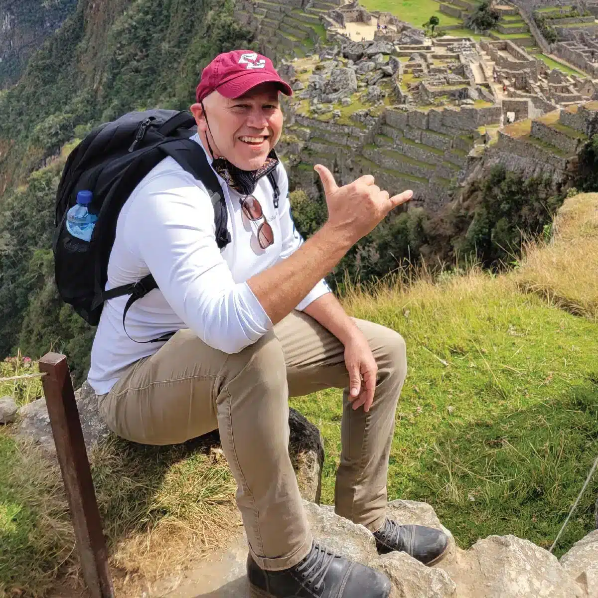 Jay McHale founder of FlowVeda sitting on a rock with a backpack giving a shaka hand sign with Machu Picchu ruins in the background