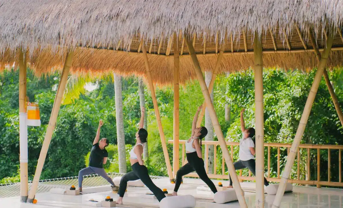 Group yoga class in an open-air thatched-roof yoga shala surrounded by tropical greenery at Goddess Retreats Bali