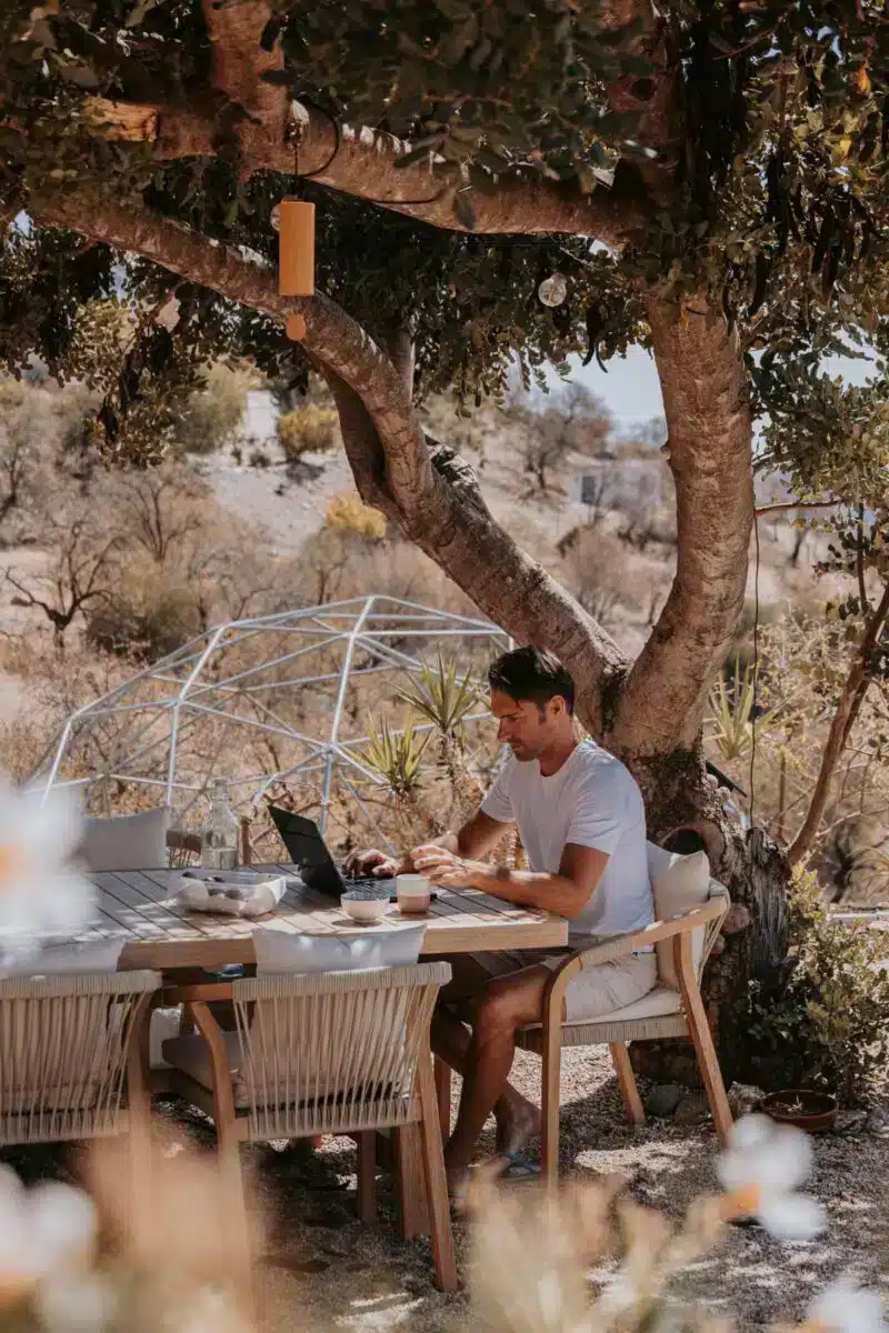 Two guests sharing a meal at a wooden table under an olive tree in the natural landscape at Costa del Soul retreat