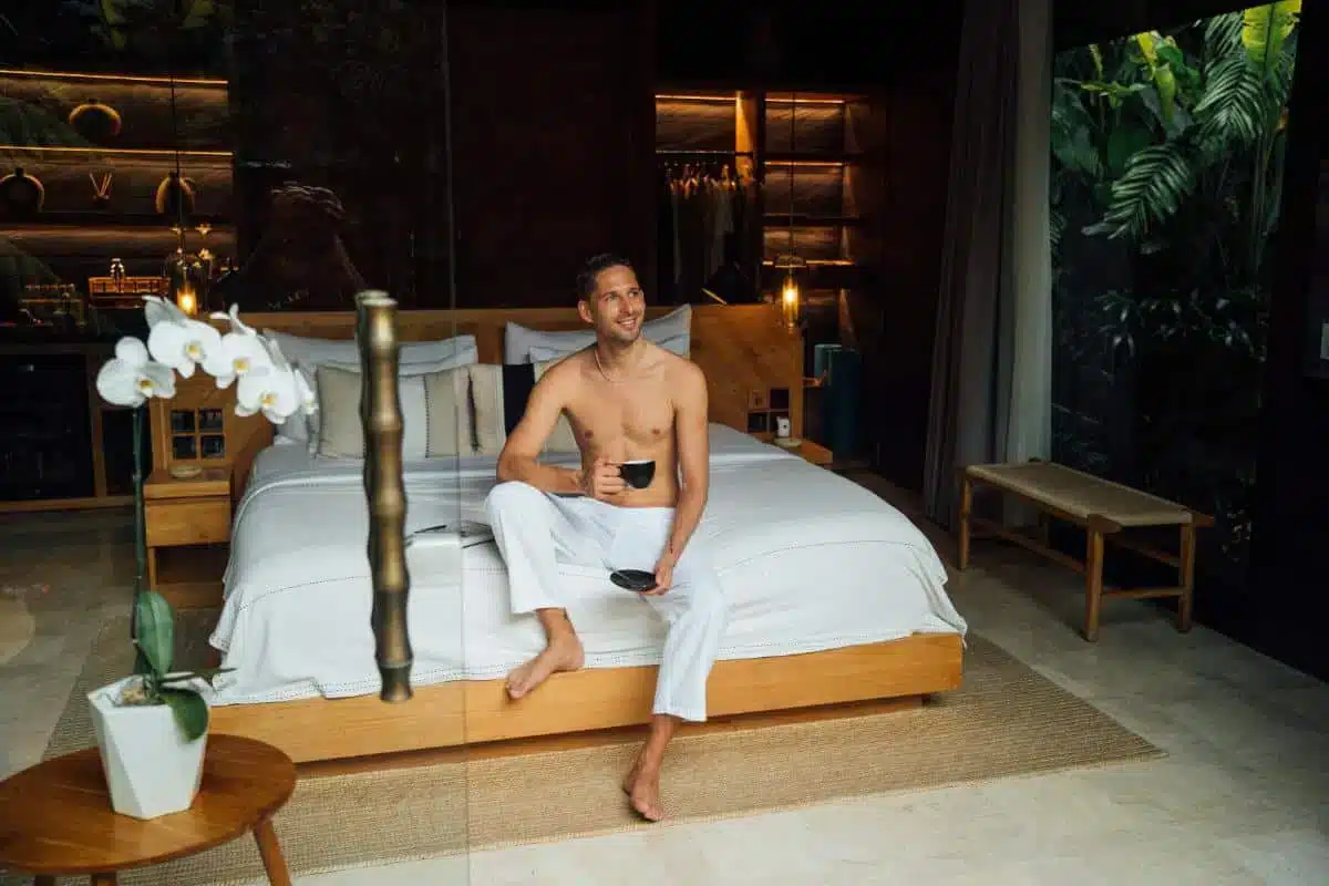 Guest sitting on the edge of the bed in the Premium Jabu Villa with wooden furniture and white orchid at Desa Hay Bali