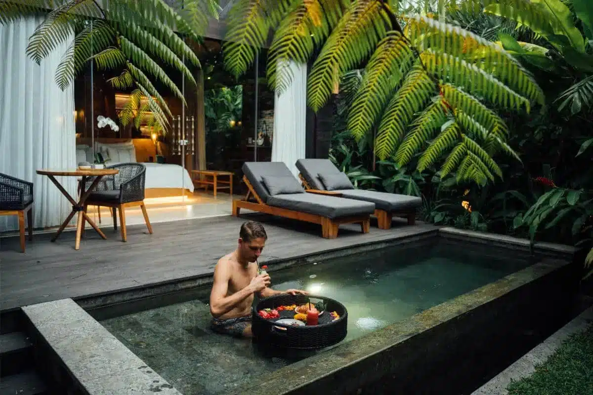 Guest sitting by the edge of the plunge pool at night with tropical garden and villa lighting in background at Desa Hay Bali