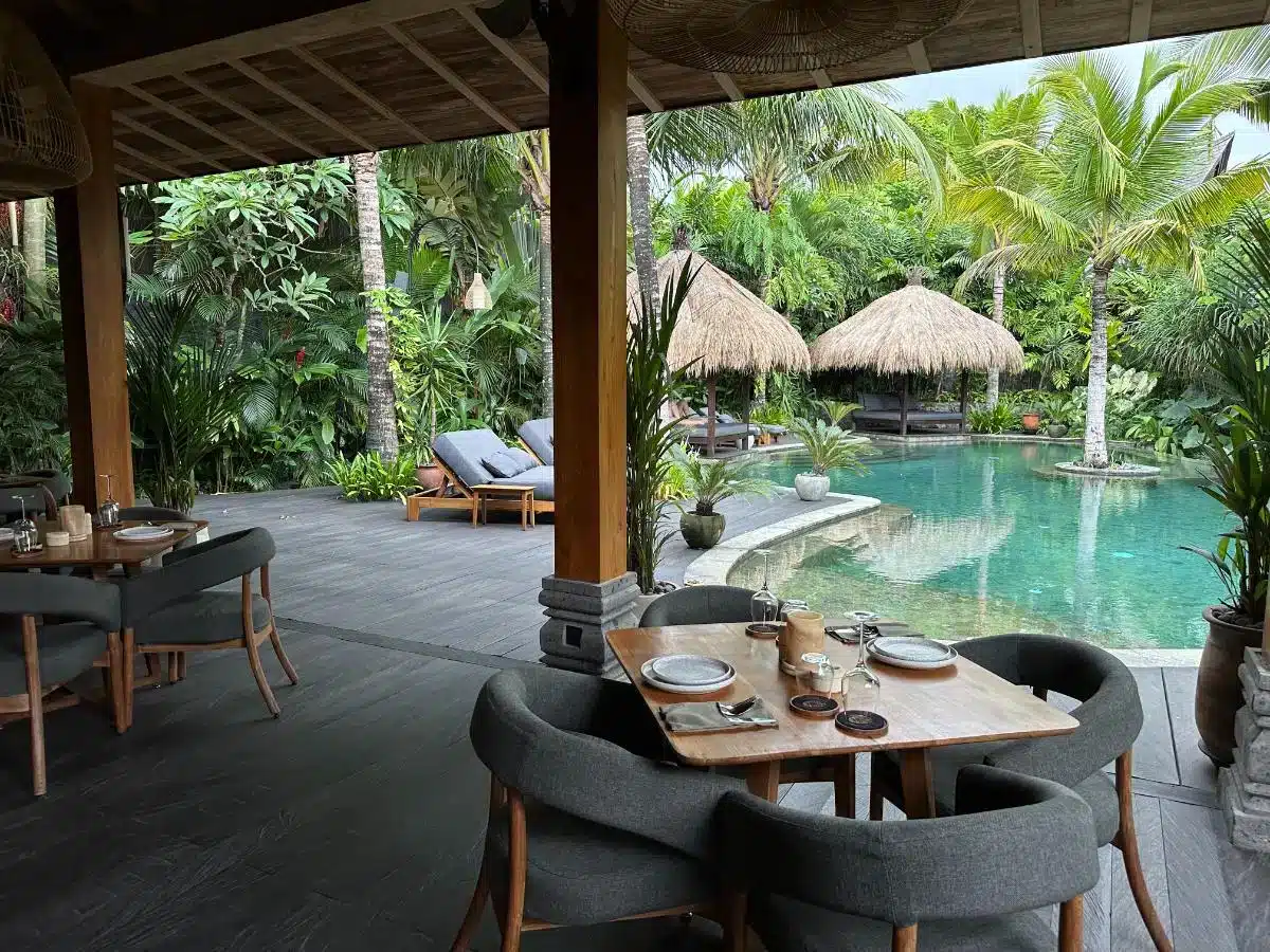 Outdoor dining area at Ijo restaurant overlooking the swimming pool with thatched umbrellas and palm trees at Desa Hay Bali