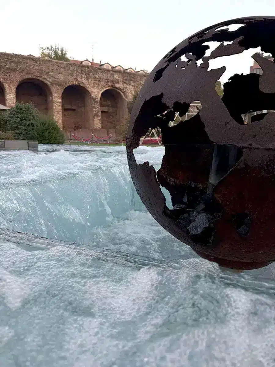 Wasser fließt durch eine Metallglobus-Skulptur mit historischen Steinbögen im Hintergrund im QC Terme Milano