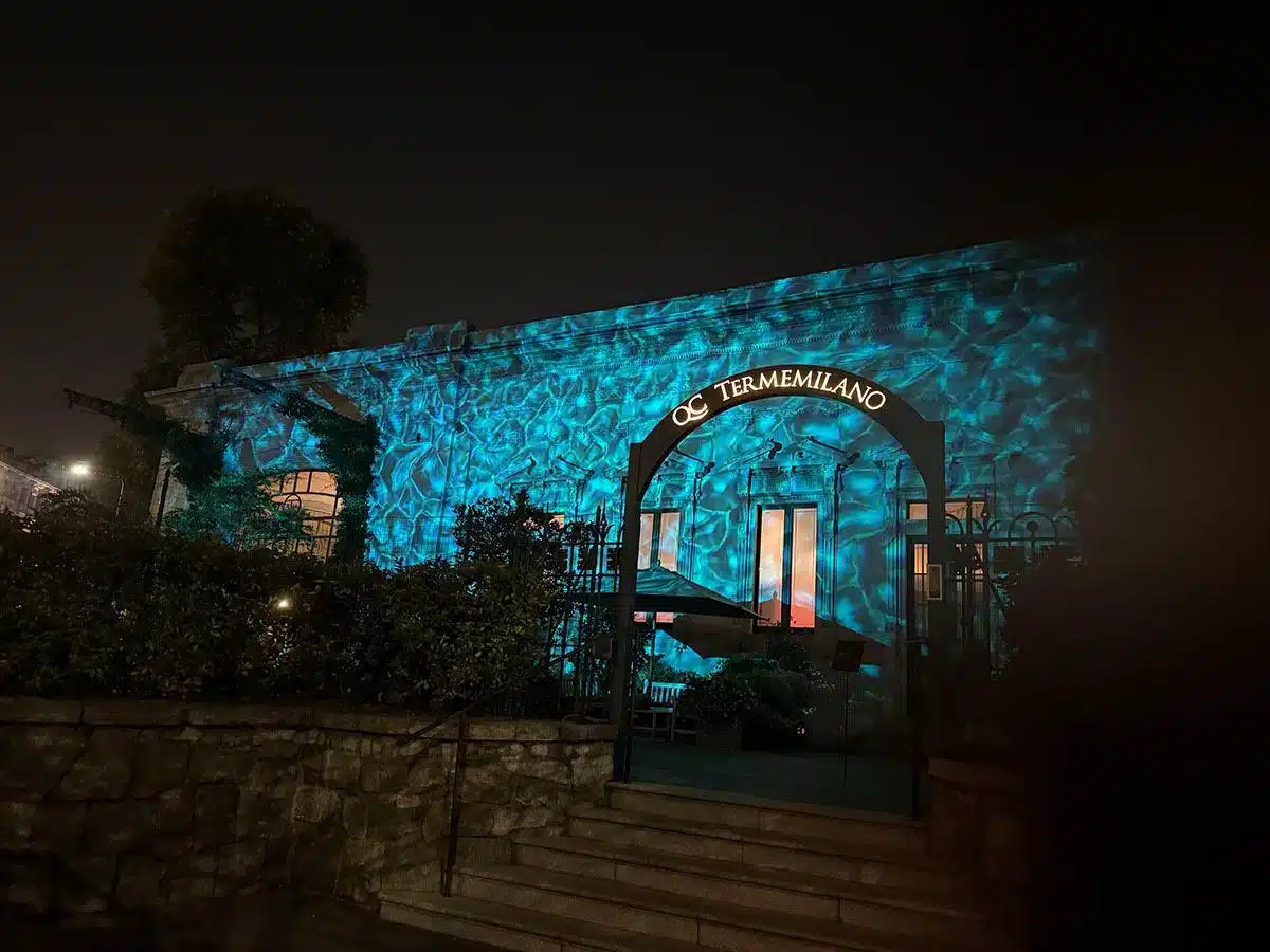 Eingangsfassade des QC Terme Milano bei Nacht mit türkisen Lichtprojektionen am Gebäude und beleuchtetem Torbogen-Schriftzug