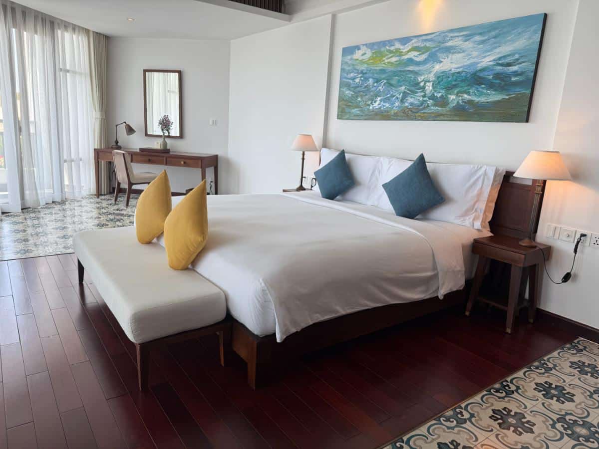Geräumiges Zimmer mit Kingsize-Bett, gelben Kissen, blauen Kunstwerken und polierten Hartholzböden im The Anam Cam Ranh