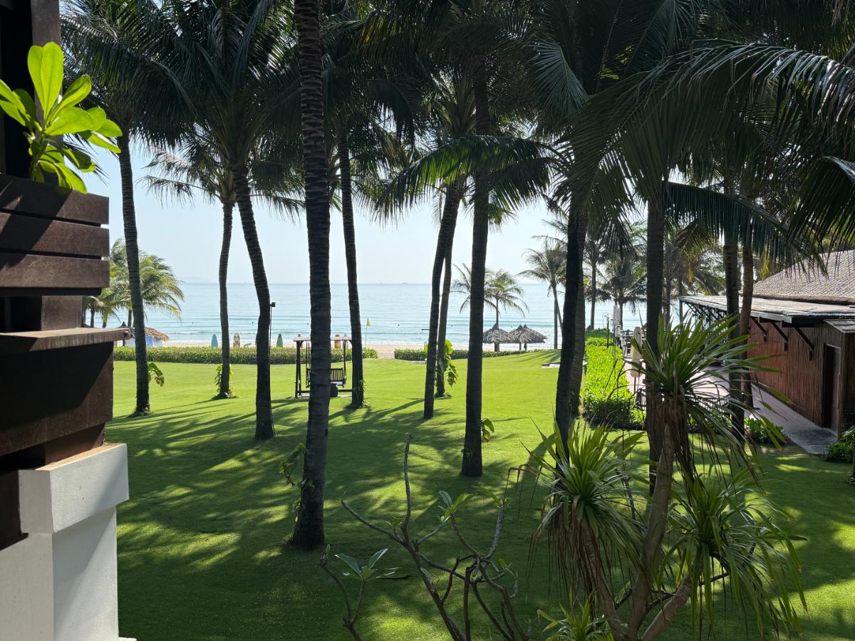 Blick vom Balkon des Zimmers auf die Gärten des Resorts, die Palmen, den grünen Rasen und das Meer im The Anam Cam Ranh
