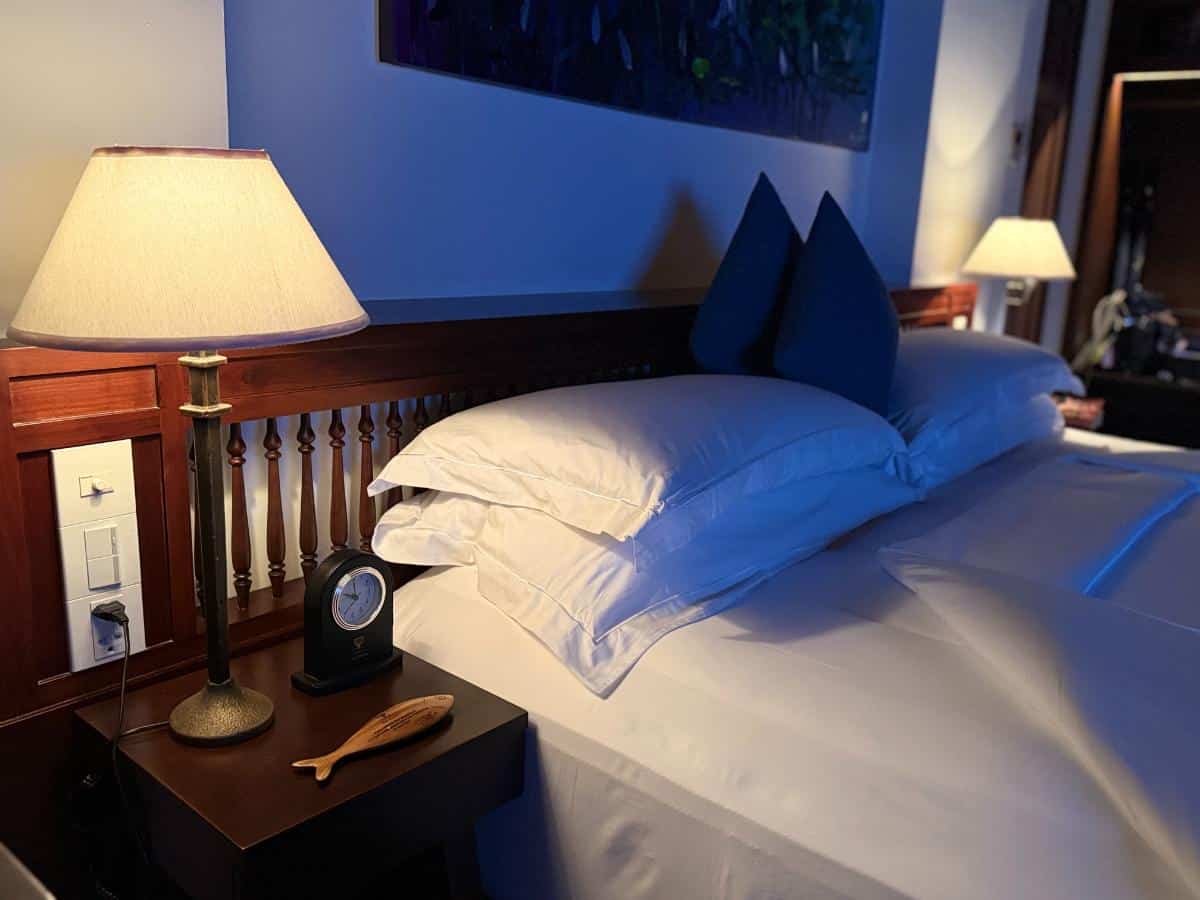 Nahaufnahme des Bettes mit frischer weißer Bettwäsche, hölzernem Kopfteil und Nachttischlampe unter der spitzen Decke im The Anam Cam Ranh