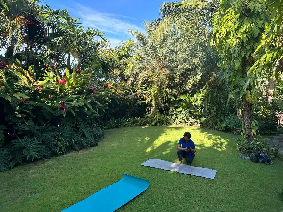 Guest practicing yoga on a blue mat on the green lawn surrounded by tropical garden and palm trees at Desa Hay Bali