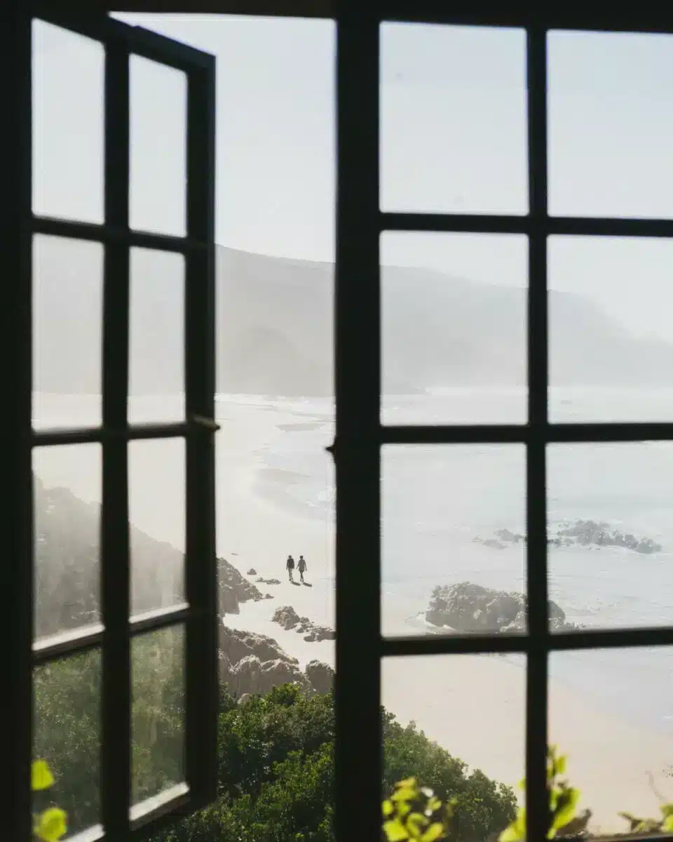 View through dark window panes of a misty coastal landscape with two silhouetted figures walking on a remote beach below, photographed at an Insider Villas property