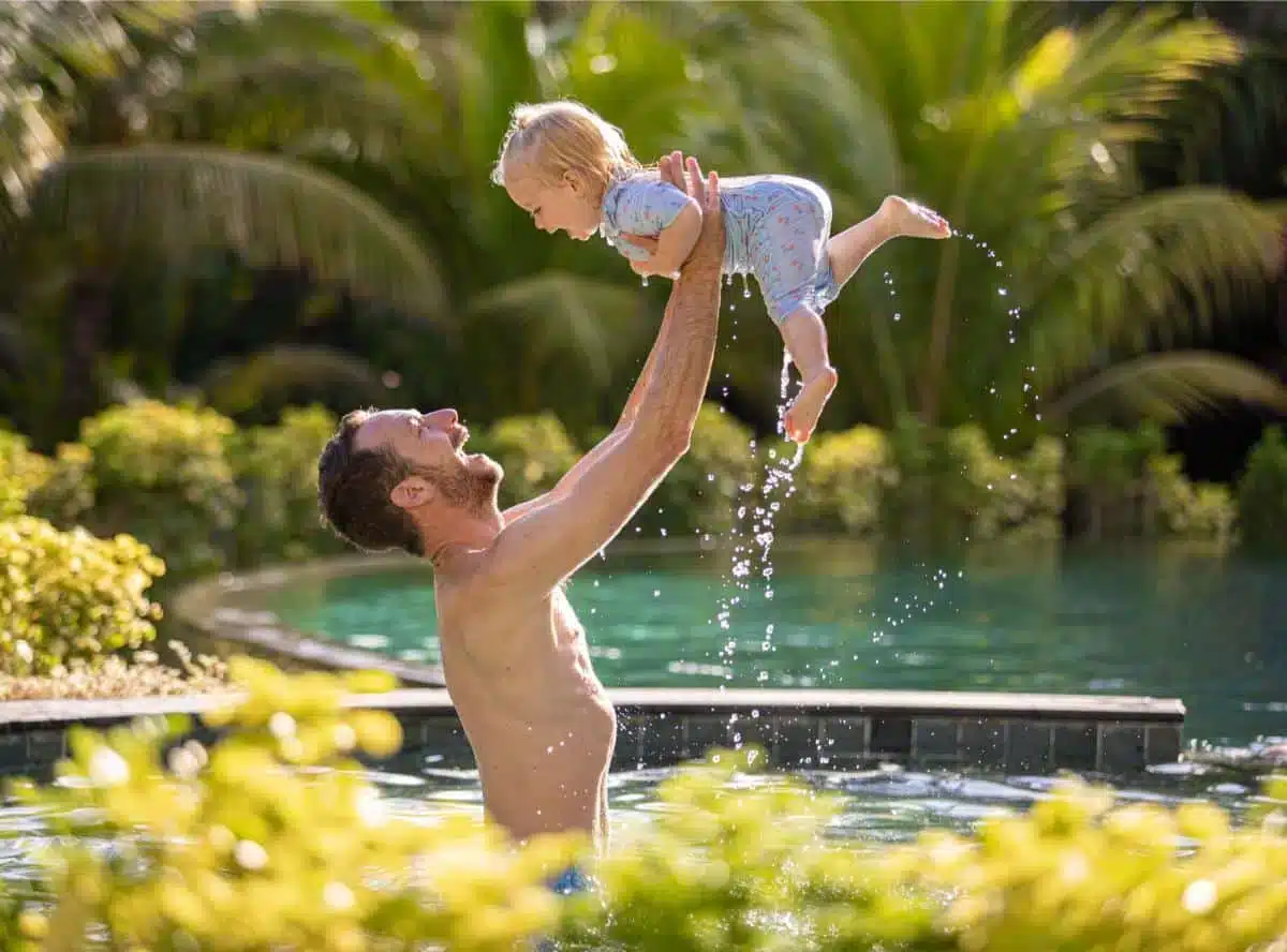 Father joyfully lifting a laughing toddler high above the water in a tropical villa pool surrounded by lush palm trees, captured at an Insider Villas property