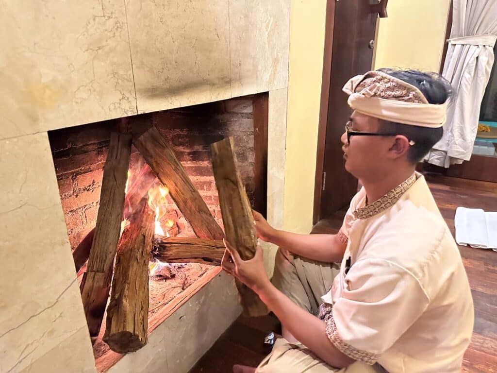 Balinese staff member in traditional attire carefully lighting kindling in villa fireplace at Munduk Moding Plantation Bali