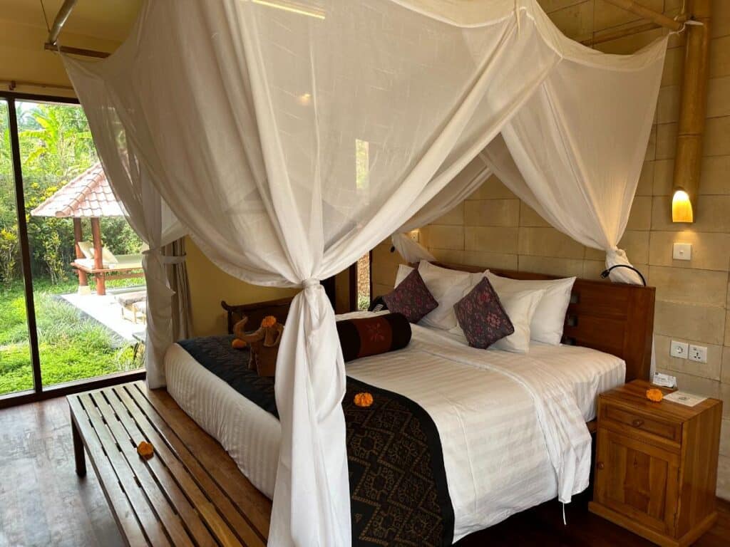 White canopy bed with sheer draping curtains in Munduk Moding Plantation villa with floor-to-ceiling windows overlooking tropical garden
