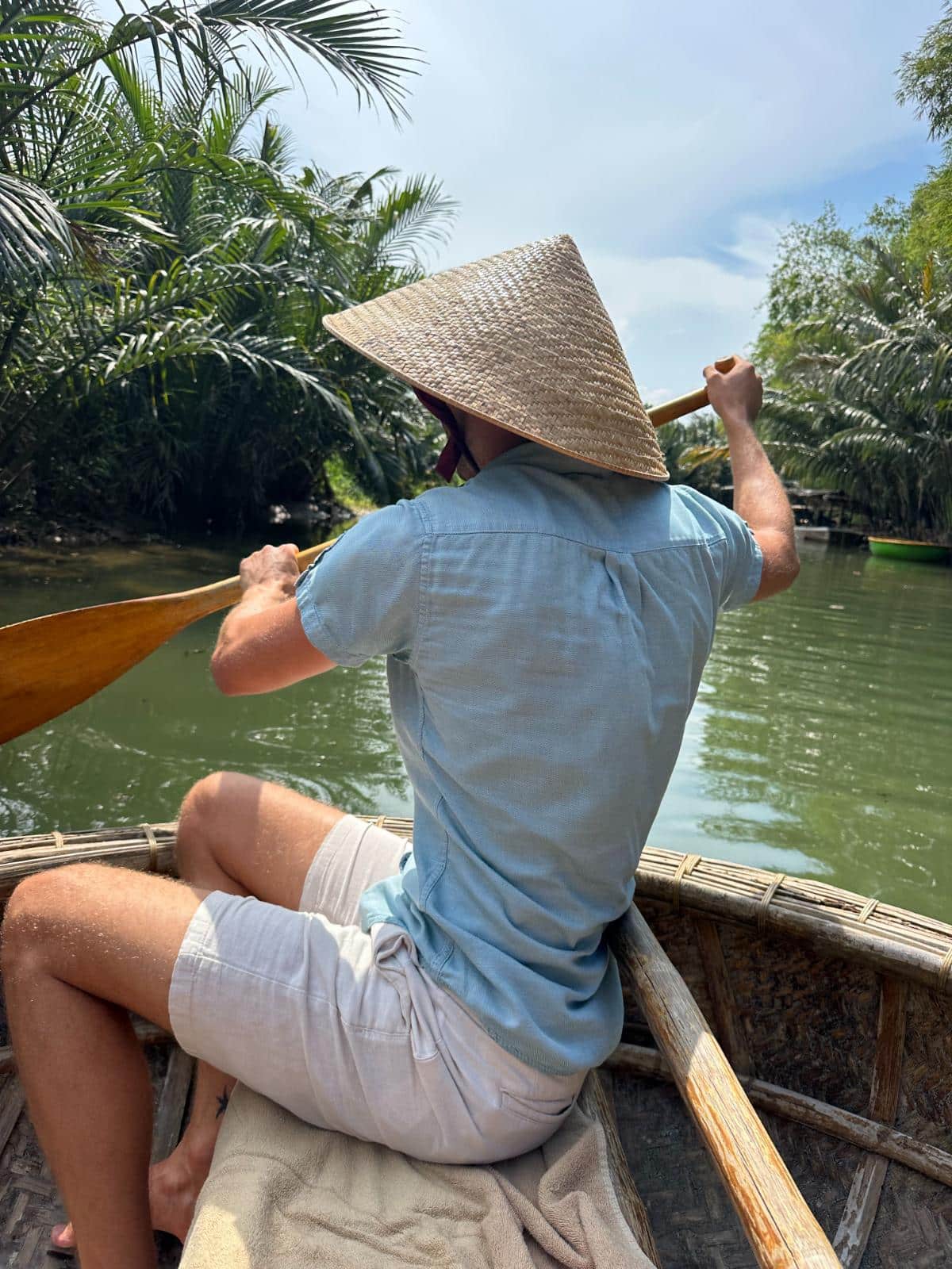Paddler mit kegelförmigem Hut steuert ein Korbboot durch die Palmenwasserstraße, Namia River Retreat, Hoi An, Vietnam