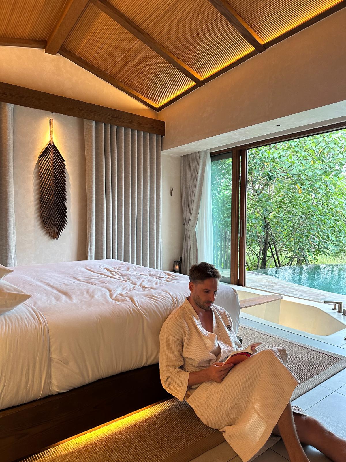 Lesen auf dem Bett am bodentiefen Fenster mit Blick auf den Pool, private Pool-Villa, Namia River Retreat, Hoi An, Vietnam